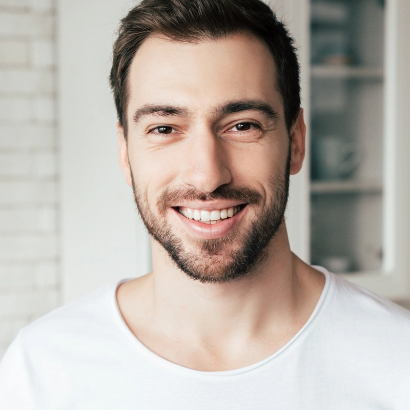 Professional headshot of smiling male dental patient in white t-shirt against blurred office background.