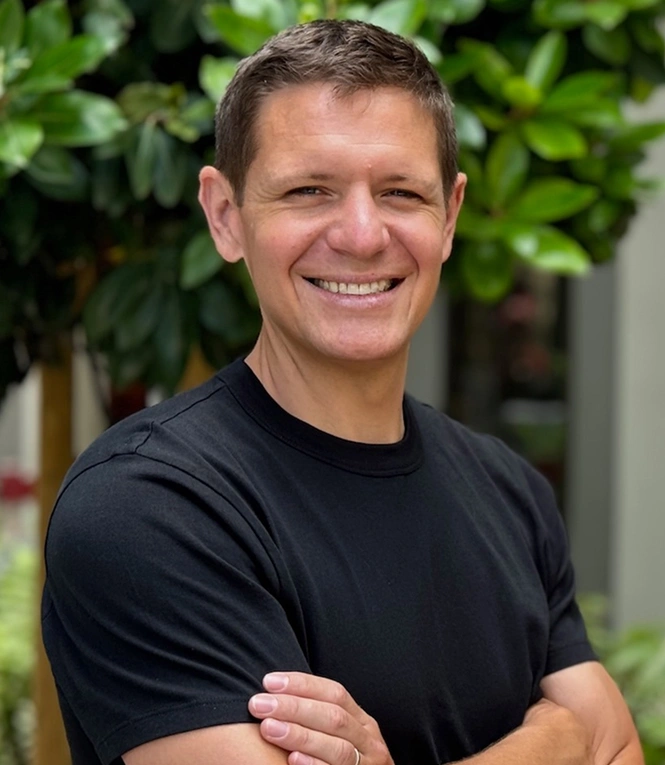 Smiling man in black shirt with arms crossed against green foliage background, professional headshot.
