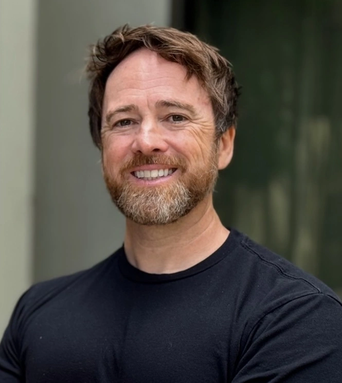 Professional headshot of male dentist smiling at camera, wearing dark shirt against blurred background.